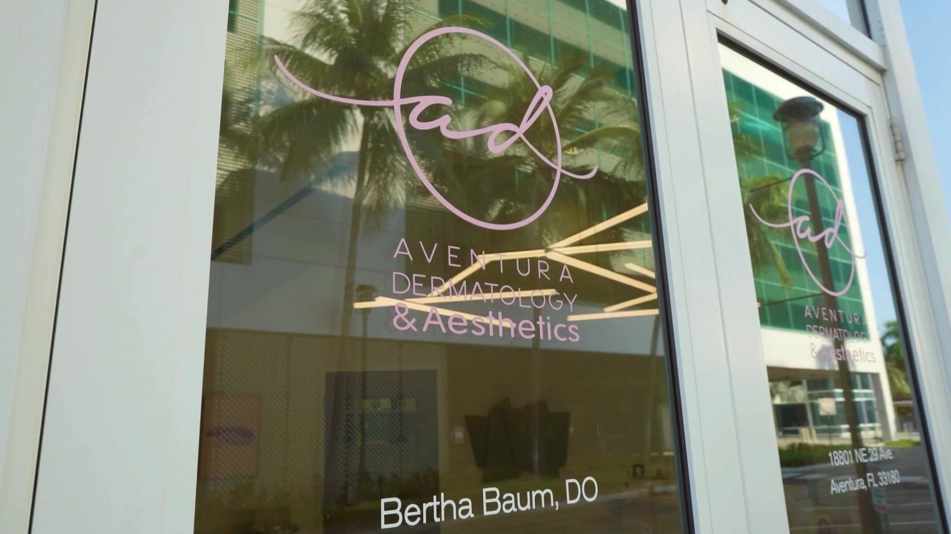 The Aventura Dermatology & Aesthetics logo and "Bertha Baum, DO" on the glass entrance doors, with palms reflected in the glass