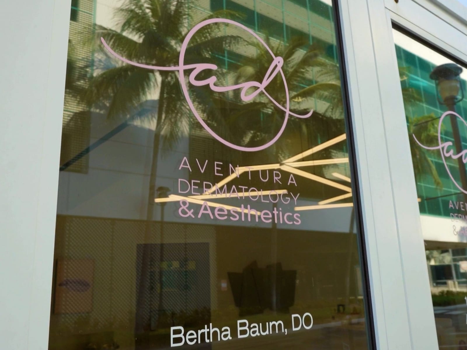 The Aventura Dermatology & Aesthetics logo and "Bertha Baum, DO" on the glass entrance doors, with palms reflected in the glass