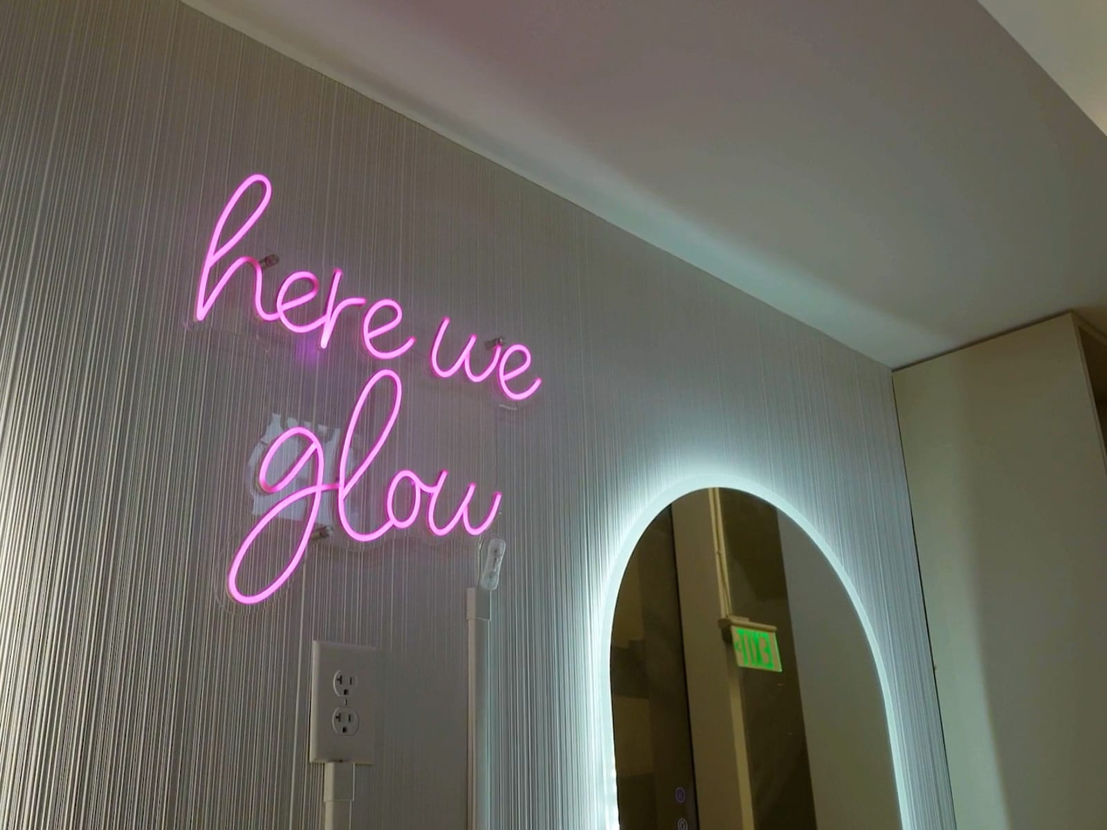 A pink neon "here we glow" sign above a backlit arched mirror
