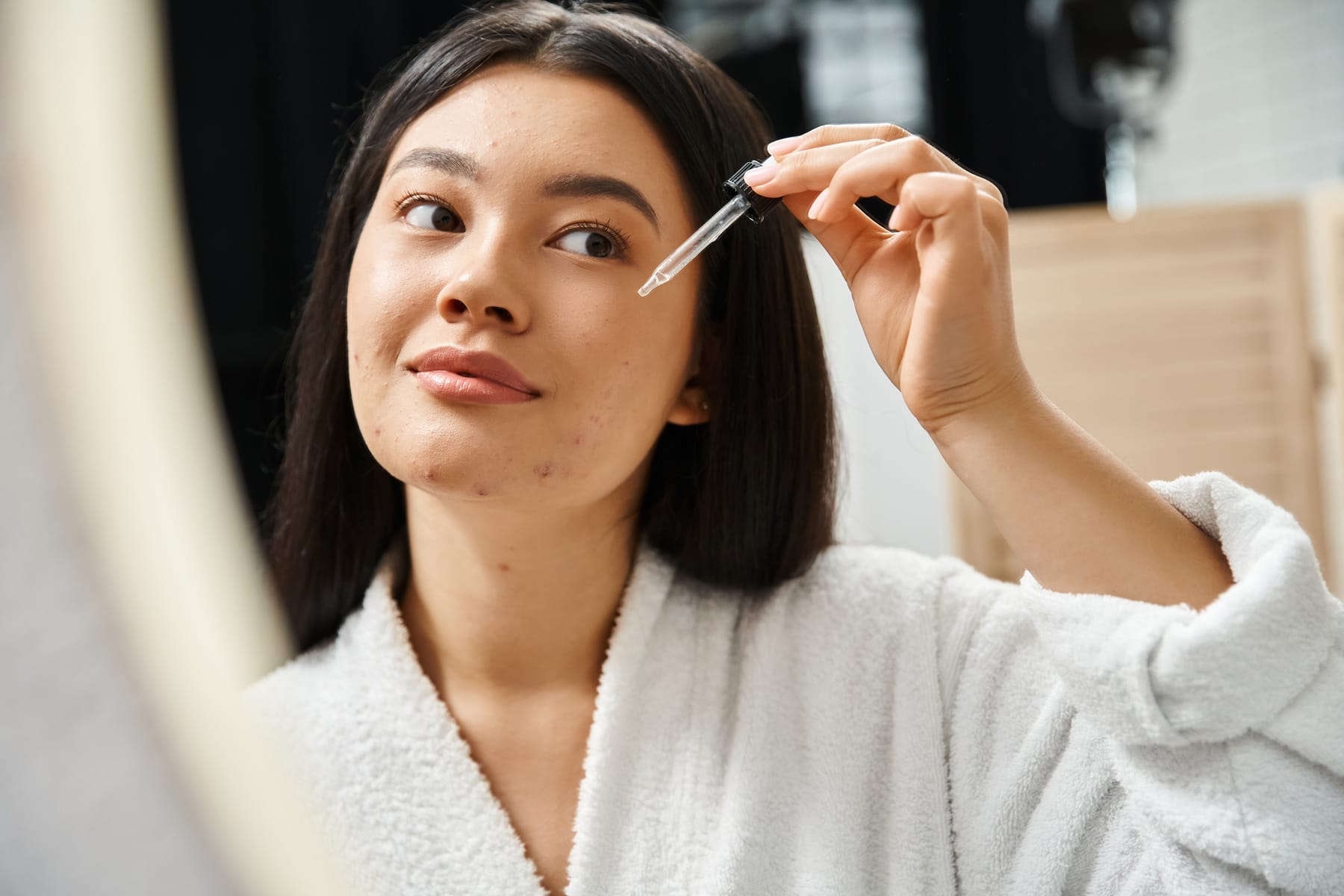 Model in a white bathrobe applying a facial serum with a dropper beside a mirror