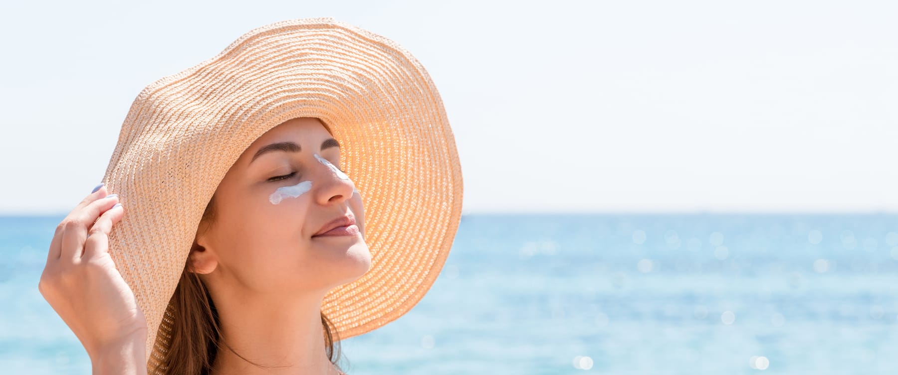 Model in a wide straw sun hat on a beach, eyes closed, with sunscreen dabbed under her eyes