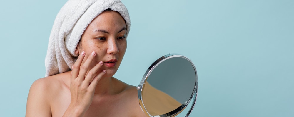 Model in a towel turban examining her skin in a hand mirror, on a pale blue background