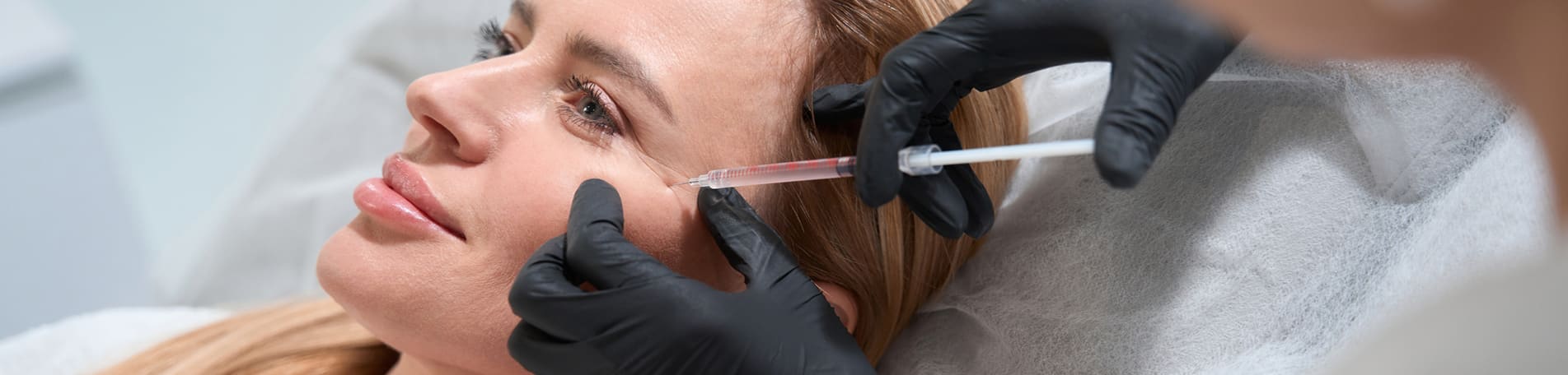 Model reclining as a clinician in black gloves injects filler beside her eye