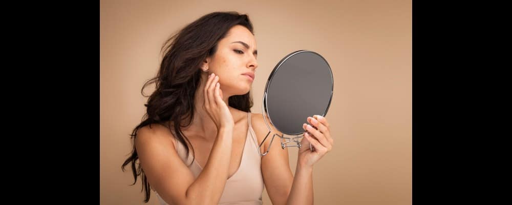 Upset woman looking at her face in the mirror, suffering with adult acne.
